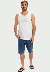White tank top with a circular logo in light green, paired with blue denim shorts and black flip-flops, on a neutral background.