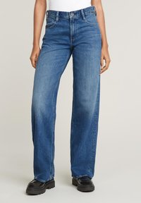 Keskmise sinise tooniga laiad denim püksid, millel on kõrge vöökoht, klassikaline viie tasku disain ja pehme pleekimine. Näha on mustad paksud platvormikingad.
