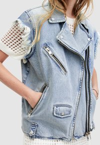 Jeansväst i ljusblått med fransade kanter, dragkedjefickor, tryckknappar och en delvis stickad vit ärmdetalj.
