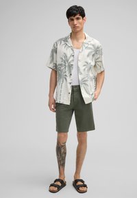 Kurzarmhemd mit Palmenmotiv, leichtes Material; kombiniert mit olivgrünen Shorts und schwarzen Slipper-Sandalen. Hautfarbene Tätowierung sichtbar.