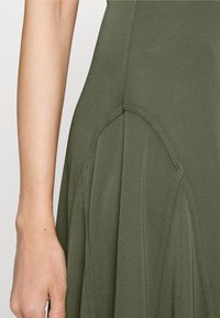 Zbliżenie na ramię i ramię modelki w oliwkowej sukience, podkreślające fakturę materiału i detale wzoru.