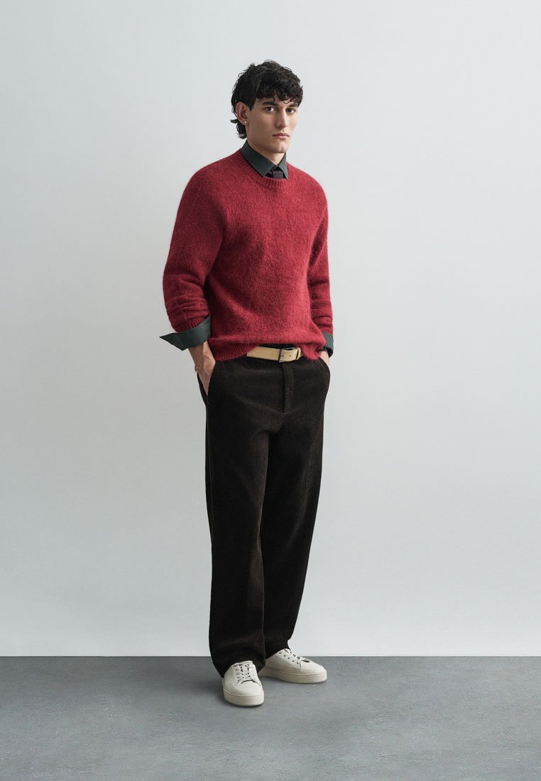 Pull en laine rouge par-dessus une chemise vert foncé, accompagné de pantalons en velours côtelé marron et de baskets blanches. Style simple, coupe décontractée avec des accents minimaux.