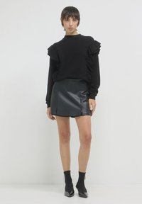 Pull noir à volants avec des manches longues, associé à une mini-jupe en cuir noir. Le mannequin porte des chaussettes noires jusqu'à la cheville et des escarpins à bout pointu.