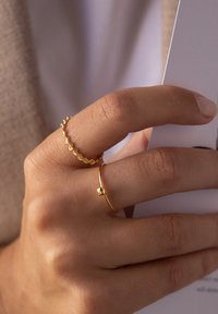 Zwei goldene Ringe an einer Hand: einer mit einem strukturierten, wellenförmigen Design und der andere ein schlichter Bandring mit einem kleinen quadratischen Akzent.