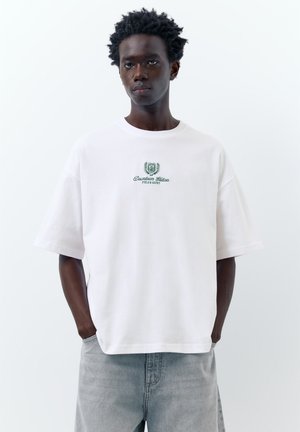 Giovane uomo con capelli scuri e ricci indossa una maglietta bianca oversize con scritta verde "Downtown Edition" e jeans grigio chiaro, mani in tasca.