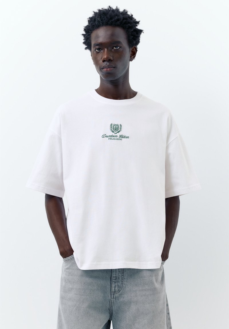 Jeune homme aux cheveux bouclés et foncés portant un T-shirt blanc oversize avec un texte vert « Downtown Edition » et un jean gris clair, mains dans les poches.