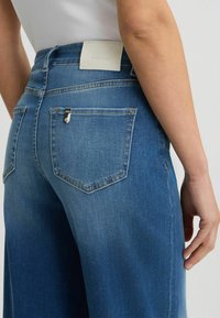 Les jeans en denim bleu à jambe large présentent une taille haute, deux poches arrière, une étiquette en cuir avec logo et un léger délavé pour ajouter de la texture.