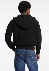 Sudadera negra de punto con textura acanalada, que presenta una capucha espaciosa y puños ajustados. Combinada con unos vaqueros de mezclilla azules que muestran un ligero desgaste.