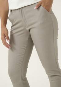 Slank gesneden, lichtgrijze broek met zijzakken, gladde textuur en subtiele naden. De stof lijkt rekbaar en figuurvolgend te zijn.