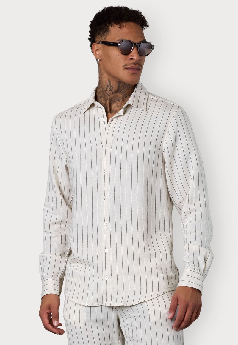 Gestreiftes Leinenhemd in Off-White mit schwarzen senkrechten Streifen, langen Ärmeln, Knopfverschluss und geradem Saum. Akzentuiert mit minimalistischem Sonnenbrille.