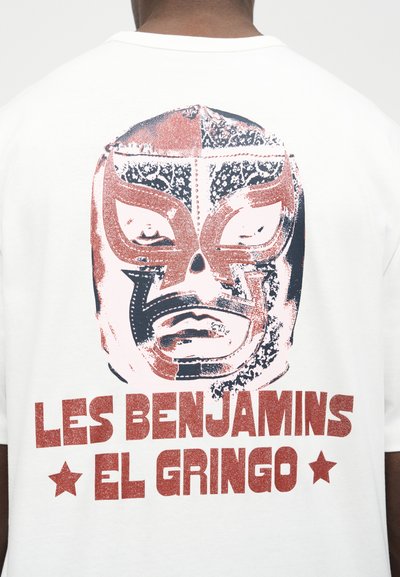 Άτομο που φοράει λευκό μπλουζάκι με γραφικό εκ stylized μάσκας πάλης σε κόκκινο και μαύρο και κείμενο «LES BENJAMINS EL GRINGO» στην πλάτη.