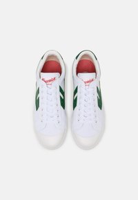White canvas sneakers with green accent stripes, round toe, lace-up design, and a red cushioned insole. Features a logo on the tongue.