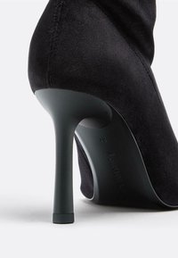 Czarny zamszowy but na wysokim obcasie o smukłym designie, z wydłużonym noskiem i błyszczącym czarnym obcasem. Zawiera subtelne detale w postaci szwów wzdłuż szwu.