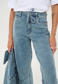 Ljusblå denimjeans med hög midja och knytdetalj. Har vida ben, fransad fåll och diskret blekt textur. Matchas med en denimjacka.