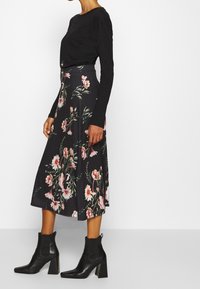 Černá dlouhá košile s dlouhým rukávem v kombinaci s květinovou sukní až po kotníky s růžovými a zelenými květy, obutá do černých espadrilek na podpatku.