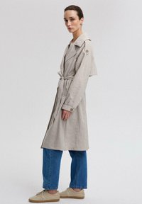 Kreemjasbeez trench coat, millel on vöökoht, suur krae ja nööbid, koos lõtvade siniste teksade ja kreemjate tossudega.