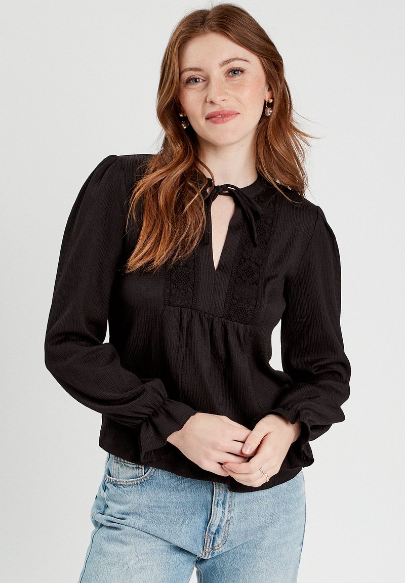 Blouse noire avec un décolleté en trou de serrure et un détail à nouer. Présente un tissu texturé, des accents en dentelle le long de l'avant et des manches bouffantes.
