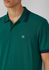 Zielona koszula polo z textured materiałem, z kontrastowym granatowym kołnierzykiem w paski oraz małym logo na piersi. Zapięcie na guziki.