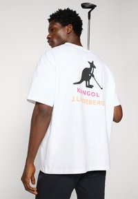 Hvid bomulds t-shirt med et sort kænguru-tryk med en golfkølle. Tekst i pink og orange siger "KANGOL J. LINDEBERG." Korte ærmer.