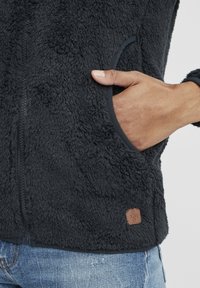 Dark gray fleece jacket with a textured surface, featuring a zip closure and side pockets. Includes a small brown logo patch on the hem.