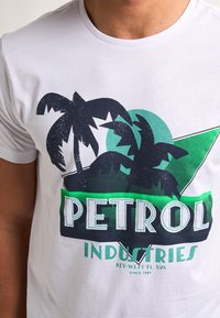 Bílý bavlněný tričko s grafikou zelené a černé palmy a textem "PETROL INDUSTRIES", představující uvolněnou designovou estetiku.