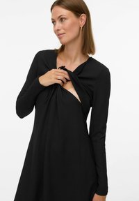 Robe noire à manches longues avec un motif texturé et un détail de nœud à l'avant révélant une ouverture partielle. Tissu lisse, corsage ajusté, jupe évasée.