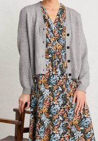 Szary, dziany kardigan z prążkowanymi mankietami i guzikami, noszony na kwiecistej sukience w zieleni, pomarańczu i błękicie. W tle drewniane krzesło.