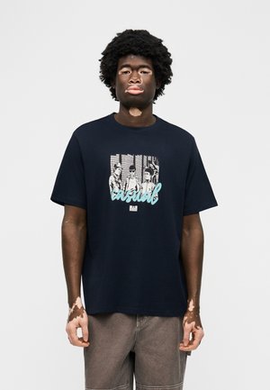 Young man with vitiligo wears a navy blue T-shirt featuring black-and-white graphic of three people and the word "casual" in light blue.