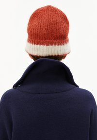Persoon met een rood-wit gebreide beanie en een marineblauwe trui met een hoge, omgeslagen kraag, van achteren gezien op een witte achtergrond.