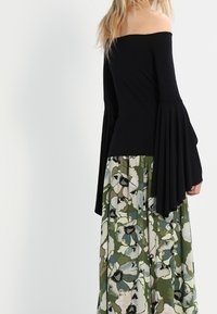 Kvinna iförd en svart off-shoulder topp med långa, utflutna ärmar och en lång grön kjol med stora vita och blå blommönster.