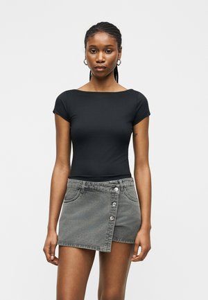 Femme aux cheveux tressés porte un haut noir ajusté à manches courtes et une mini-jupe en jean grise asymétrique avec des boutons à l'avant sur fond blanc.