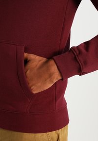 Sudadera de color marrón con un bolsillo canguro y puños acanalados. La tela parece suave y texturizada, con un ajuste clásico y un diseño sin costuras.