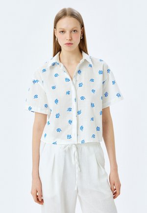 Camisa blanca de manga corta con patrones florales azules, con botones en el frente y cuello. Combinada con pantalones blancos holgados, hechos de una tela ligera.