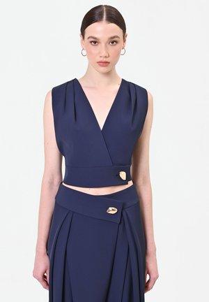 Donna che indossa un crop top senza maniche blu navy e gonna a vita alta abbinata con bottoni decorativi dorati su sfondo bianco.