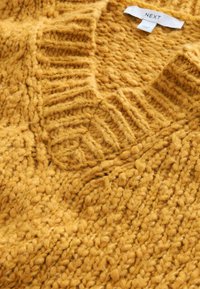 Sennepsgul strikket sweater med en bred v-udskæring, der har tekstureret ribstruktur og et generelt chunky, fuzzy design. Mærke synligt indeni.