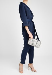 Žena v tmavě modrém overalovém oblečení s páskem, držící šedou kabelku se vzorem hadí kůže, oblečená v stříbrných vysokých podpatcích a s náramkem.