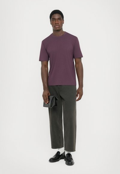Man wearing purple t-shirt, black cropped pants, white socks, black loafers, holding a black clutch against a plain light background.