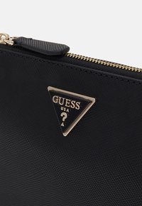Mustalla tekopinnalla varustettu vetoketjullinen pussi, jossa on kultaiset yksityiskohdat ja kolmiomainen logo-plaakkia, jossa lukee "GUESS USA" ja kysymysmerkkiyksityiskohta.