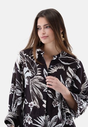 Junge Frau mit langen braunen Haaren, die goldene Ohrringe trägt und ein schwarzes Hemd mit weißen tropischen Blatt- und Blumenmustern, hält einen Knopf in der Hand.