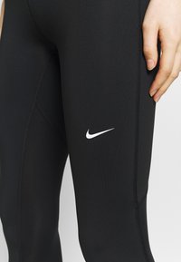 Czarne legginsy wykonane z elastycznego materiału, z białym logo Nike po lewej stronie i widocznymi szwami wzdłuż krawędzi.