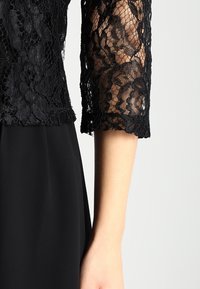 Svart spetsblus med trekvarts ärm som visar intrikata blommönster över den bara armen, ihop med en enfärgad svart kjol eller klänning.