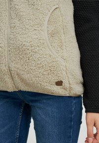 Beige Fleece-Weste aus strukturiertem Stoff, mit Seitentaschen und dunklen Strickärmeln. Verfügt über ein kleines braunes Logopatch am Saum.