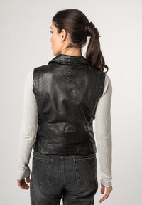 Gilet in pelle nera con colletto e dettagli di cucitura verticale. Indossato sopra una camicia a maniche lunghe grigia chiara, abbinato a jeans scuri.