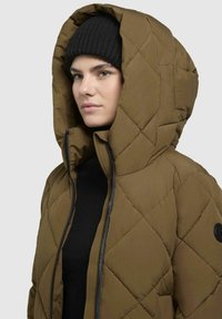 Brown quilted puffer jacket with a large hood, featuring a zipper closure and black accents. Worn over a black sweater, with a knit beanie.