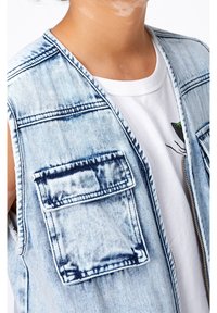 Džínová vesta ve světle modré barvě, bez rukávů s dvěma náprsními kapsami. Texturovaná tkanina s viditelným prošíváním a detaily opotřebení. Pod ní bílý tričko.