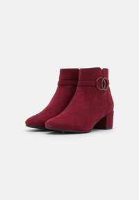 Bottines en daim bordeaux avec un talon bloc, dotées d'une sangle décorative et d'un accent en métal circulaire sur le côté. Texture lisse.
