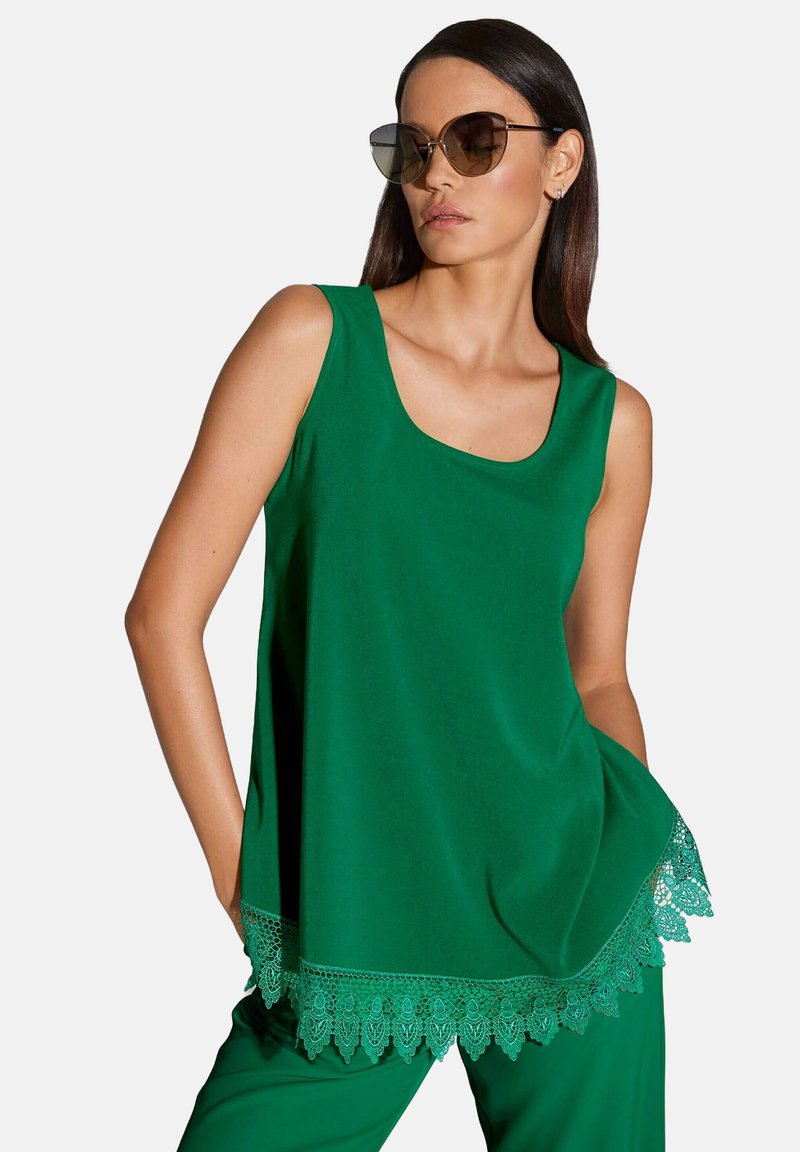 Grünes ärmelloses Oberteil aus leichtem Stoff mit rundem Halsausschnitt und kunstvollem Spitzenbesatz am Saum. Kombiniert mit grünen Hosen.