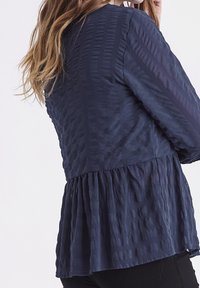 Námořnická blůza s peplum střihem, vyrobená z texturované tkaniny, s vertikálními pruhy a nabíraným lemem. Dlouhé rukávy s jemným leskem.