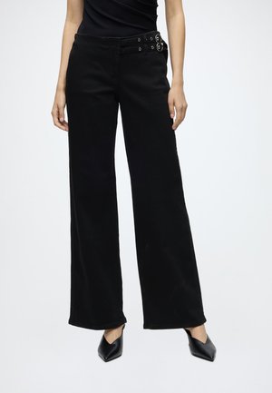 Femme portant un pantalon noir taille haute à jambes larges avec un détail de double boucle à la ceinture et des talons noirs pointus, debout sur un fond blanc.