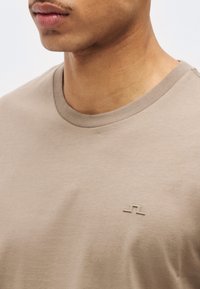 Κοντινό πλάνο ενός ατόμου που φοράει μια μπεζ κοντομάνικη μπλούζα με στρογγυλή λαιμόκοψη και ένα μικρό, ανάγλυφο λογότυπο στο αριστερό μέρος του στήθους.